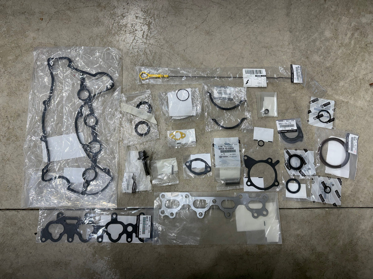 Mazda OEM engine gasket set for the NA/NB Miata laid out on the shop floor: valve cover gasket, intake and exhaust manifold gaskets, front and rear main seals, oil pan gaskets, dipstick, water pump gasket and the other seals in their Mazda bags