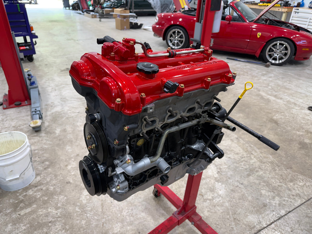 Assembled Miata engine with red valve cover and yellow dipstick handle on a red engine stand