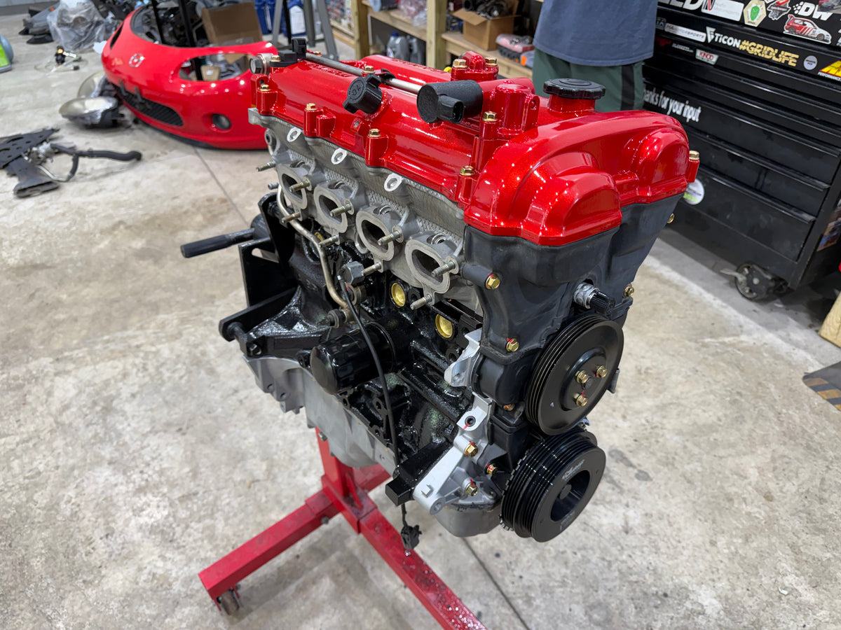 Assembled Miata engine with red valve cover and black front pulleys on a red engine stand