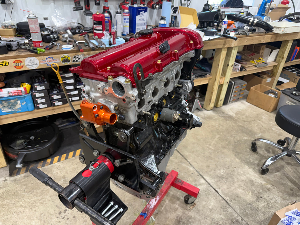 Assembled Miata engine with red valve cover and orange coolant outlet on an engine stand, rear view