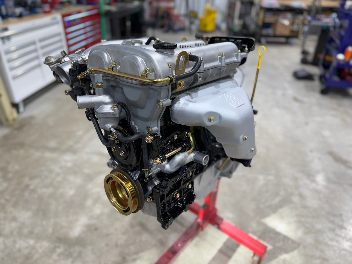 Assembled Miata engine on a build stand with silver valve cover, exhaust heat shield and gold crank pulley
