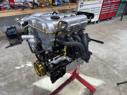 Assembled Miata engine with silver valve cover, black exhaust header and gold crank pulley on a red engine stand